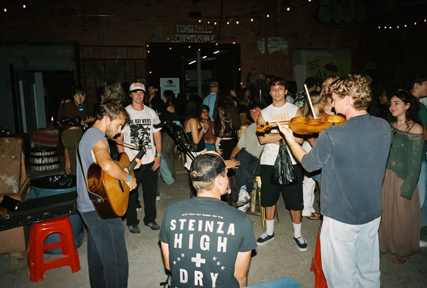 Two musicians playing guitar and fiddle in the middle of the crowd at the Palm Radio Launch Party